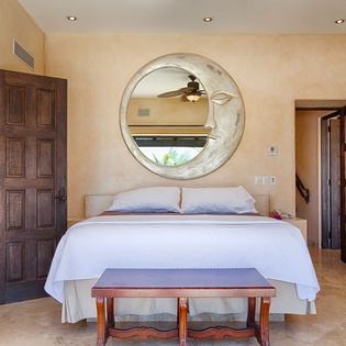 Your serene bedroom features a plush bed beneath a striking circular mirror, warm tile floors, and natural light filtering through—your perfect retreat for restful nights.
