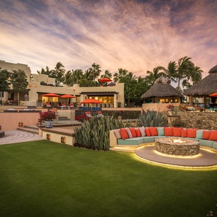 Stunning multi-level resort property with curved seating area, tropical landscaping, and traditional thatched-roof structures set against a dramatic sunset sky.