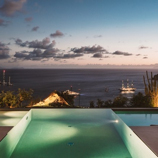 Illuminated infinity pool overlooking dramatic ocean views with cruise ships dotting the horizon at dusk.