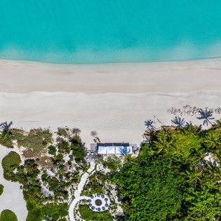 Aerial view captures a beachfront property nestled between lush tropical vegetation and pristine white sand meeting crystalline turquoise waters.