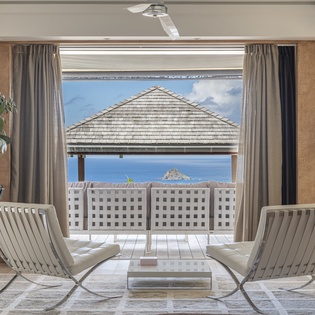 Relax in this serene lounge with elegant seating and sweeping views through an open balcony door, framing brilliant blue waters and distant islands beyond.