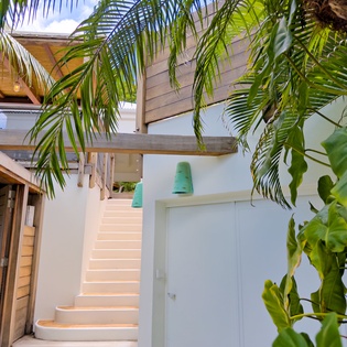 Lush tropical foliage frames the charming outdoor stairway leading to your private upper-level retreat, blending modern coastal design with natural island beauty.