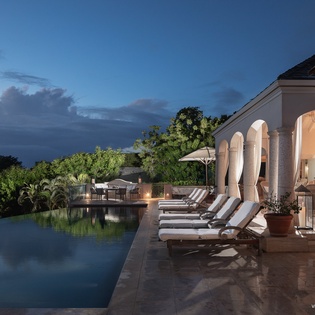 Relax poolside as the day fades, with elegant arches framing your illuminated living space and lush tropical trees surrounding the infinity pool at twilight.