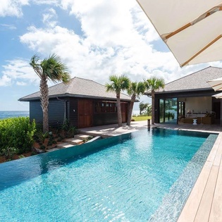 Oceanfront infinity pool with stunning sea views and tropical landscaping, surrounded by comfortable lounge seating and palm trees. Modern property architecture frames this private retreat.