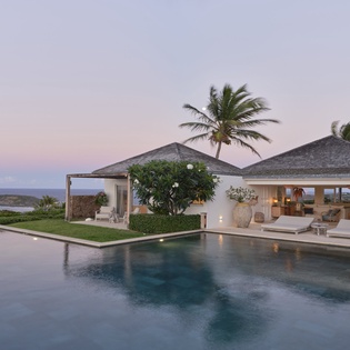 A stunning oceanfront villa bathed in golden dusk light, with a tranquil infinity pool reflecting the soft sky and elegant palm trees swaying in the breeze—pure island serenity.
