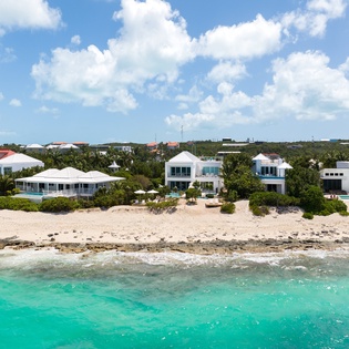 Stunning beachfront properties line pristine turquoise waters and white sand beach under bright blue skies.