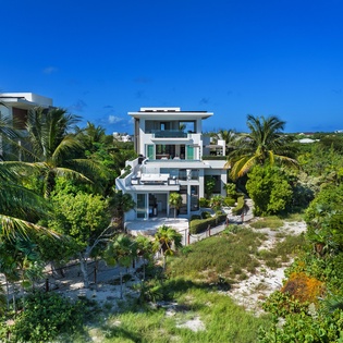 Modern beachfront villa nestled among tropical palms and lush vegetation under brilliant blue Caribbean skies.