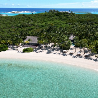Stunning aerial view reveals a pristine tropical paradise with crystal-clear turquoise waters, powdery white sand beach, and lush palm grove surrounding traditional thatched-roof buildings.