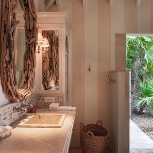 Step into a sun-lit sanctuary where natural driftwood mirrors frame your vanity and an open door invites tropical breezes. This spa-inspired bathroom blends earthy textures with serene garden views for your perfect escape.