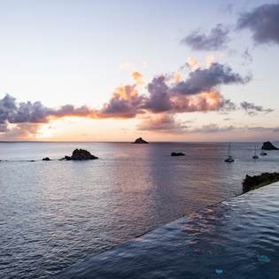 Infinity pool overlooks dramatic coastline as sunset paints the sky in brilliant hues over scattered islands and anchored sailboats.
