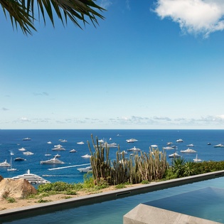 Infinity pool overlooking crystal-clear Mediterranean waters dotted with luxury yachts, framed by tropical gardens and distant island peaks.