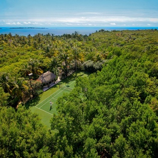 Aerial view of tropical property with tennis court nestled in lush jungle landscape near pristine coastline.