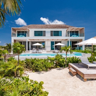 Modern beachfront villa with pristine pool surrounded by tropical palms and white sand.
