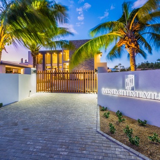 The illuminated entrance gate and property sign welcome guests to Wind Chime Villa at dusk, framed by swaying palms and a peaceful tropical setting.