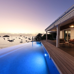 Stunning beachfront property featuring an infinity pool overlooking the harbor at sunset, with elegant outdoor seating areas.