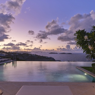 An infinity pool reflecting pastel skies and overlooking tropical islands. Lush greenery frames the peaceful scene as day transitions to evening.