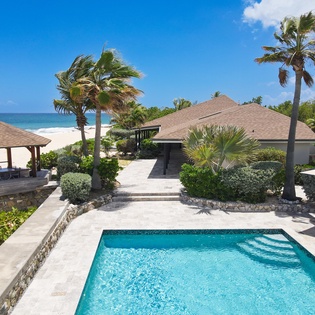 A stunning beachfront property featuring a pristine private pool, shaded gazebo, and swaying palms set against turquoise waters and soft white sand—luxury coastal living at its finest.