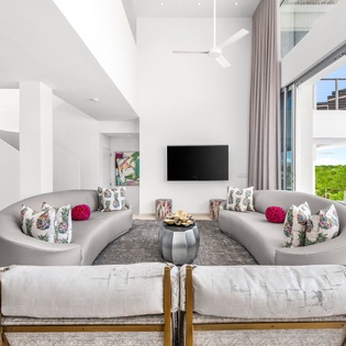 Relax in this stunning open-concept living space with curved sofas, soaring ceilings, and floor-to-ceiling windows framing tropical views.