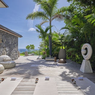 The property's deck blends stone-and-wood architecture with tropical palms, modern sculpture, and lounge seating overlooking the sea—a tranquil outdoor retreat in a lush natural setting.