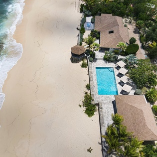 Aerial view of a beachfront villa featuring a private pool, lush tropical gardens, and direct access to pristine white sands and turquoise waters.