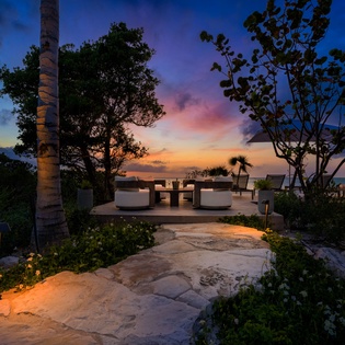 Stunning sunset paints the sky in vibrant hues over the oceanfront terrace, framed by lush tropical landscaping and inviting seating for evening relaxation.