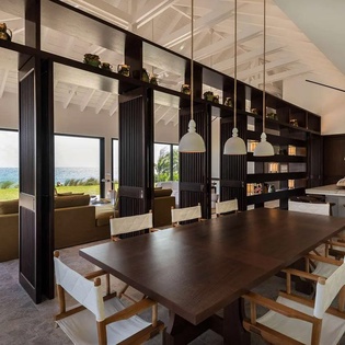 Step into your open-concept living space where a grand dining table anchors the room under striking pendant lights. Natural light pours through floor-to-ceiling windows framing stunning ocean views, while modern furnishings and custom shelving create the p