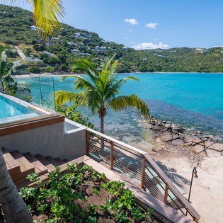 Stunning waterfront property features infinity pool overlooking pristine turquoise bay with tropical palms and hillside homes dotting the lush landscape.