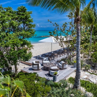 Private beachfront deck framed by palms and lush tropical foliage, with modern seating overlooking pristine white sand and turquoise waters—a secluded slice of paradise.