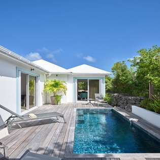 Modern white villa with private swimming pool surrounded by wooden decking and lush tropical landscaping under clear blue skies.