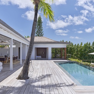Stunning tropical villa features a pristine pool deck beneath swaying palms, with mountains and lush gardens framing the property, offering an idyllic Caribbean escape.