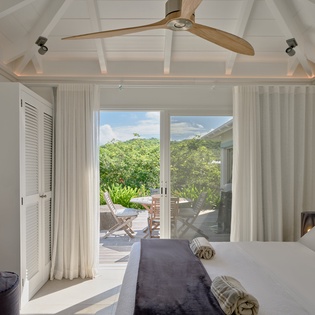 Relax in your serene bedroom where vaulted ceilings and flowing white curtains frame tropical garden views through expansive doors.