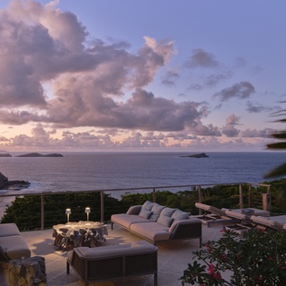 Twilight paints the sky above a serene beachfront terrace overlooking tropical islands and a quiet cove. Property exterior at dusk with ocean and surrounding landscape.