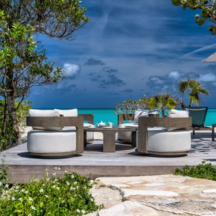 A serene beachfront terrace with cushioned seating overlooks brilliant turquoise waters, framed by tropical palms and lush greenery—your perfect spot to unwind in paradise.