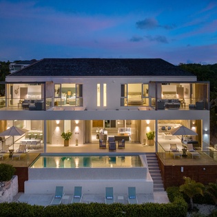 Modern luxury villa with illuminated interiors and private pool, nestled among tropical vegetation in an exclusive island setting.