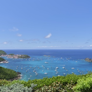 Stunning coastal landscape featuring a lush hillside overlooking a vibrant turquoise bay dotted with sailboats, with distant islands and clear blue skies visible on the horizon.