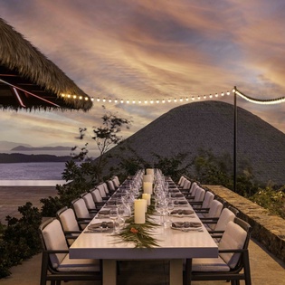 Elegant beachside dining pavilion set for an evening celebration, featuring thatched roofs and ocean views under a stunning tropical sunset.