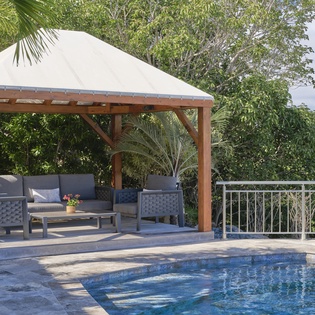 Relax under your private poolside gazebo, where comfortable seating awaits beside the sparkling blue water surrounded by lush tropical gardens.