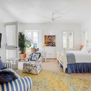 Picture yourself unwinding in this bright coastal bedroom, where soft linens, a vibrant area rug, and comfortable seating create your perfect retreat. The ceiling fan keeps you cool while you enjoy ocean breezes through shuttered doors.