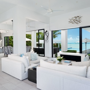 Sink into plush white sofas while turquoise waters and swaying palms frame your perfect tropical escape through floor-to-ceiling windows.