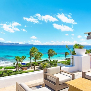Beachfront terrace with stunning turquoise waters and mountain views, framed by swaying palms and modern outdoor seating perfect for seaside relaxation.