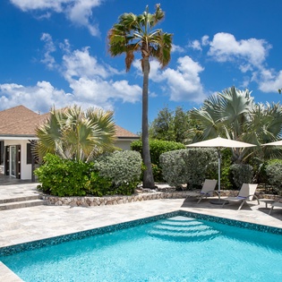 A stunning private retreat with sparkling blue pool, sun loungers beneath shaded umbrellas, and lush tropical landscaping under bright Caribbean skies.