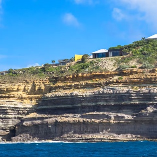 A striking hillside property nestled above dramatic coastal cliffs with layered sedimentary rock formations, lush greenery, and vibrant blue waters framing the scenic surrounding area.