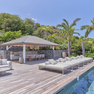 Luxurious poolside paradise with pristine swimming pool, elegant lounge beds, covered dining pavilion, and lush tropical landscaping.