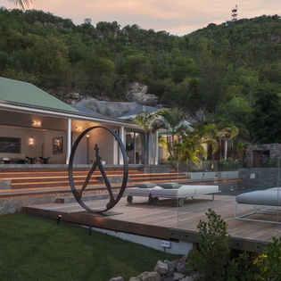 Modern villa nestled in lush tropical hills, featuring contemporary design with glass walls and sculptural elements at twilight.