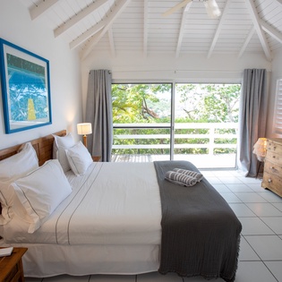 Your serene bedroom features exposed white beams, crisp linens, and a peaceful garden view through sliding doors onto your private balcony.