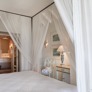 Drift into dreamy sleep beneath billowing white canopy drapes. This serene bedroom welcomes you with soft linens, elegant lamps, and a charming en-suite just steps away.