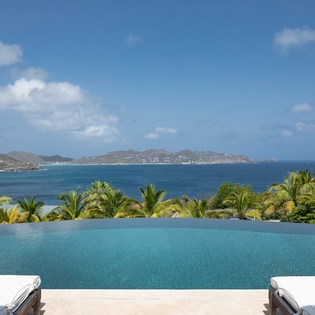 An infinity pool frames sweeping views of turquoise waters, lush palms, and distant coastal mountains under open Caribbean skies.