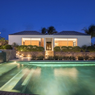 Modern luxury villa with illuminated swimming pool and tropical landscaping under evening sky.