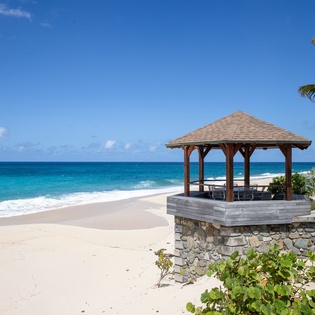 A covered gazebo with seating overlooks pristine white sands and turquoise waters, framed by swaying palms and tropical blooms—a perfect beachfront retreat.