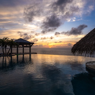Overwater bungalow with thatched roof overlooks tranquil lagoon waters as golden sunset paints the sky in warm tropical hues.